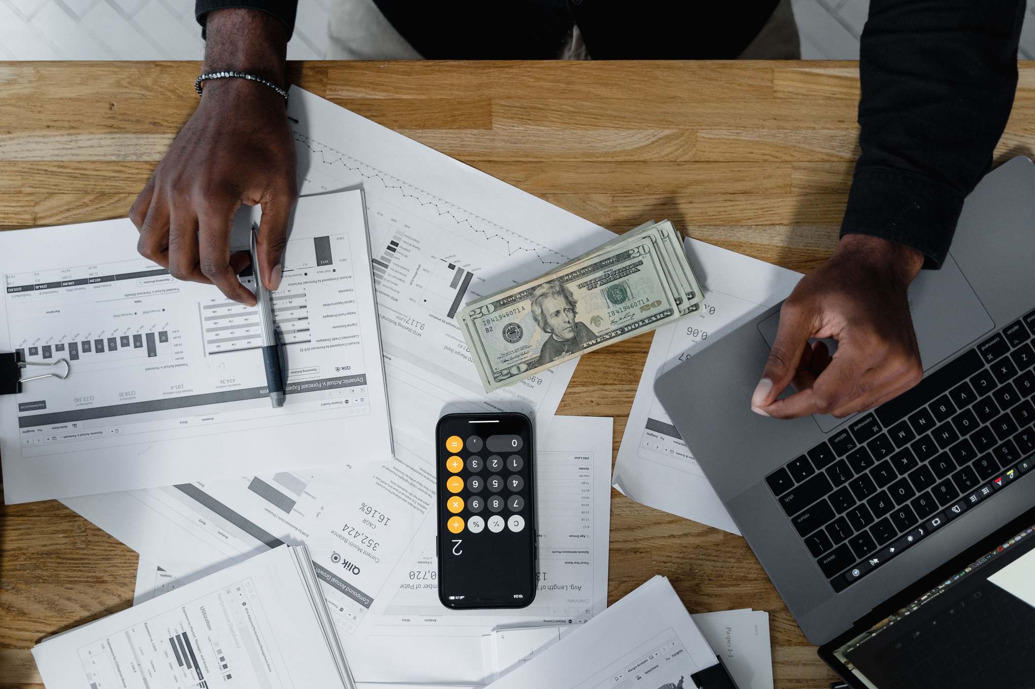 Hands calculate finances with papers, cash, and a laptop on a wooden desk